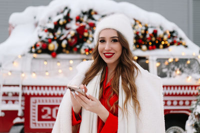 A beautiful young woman with red lips in warm hat stands by a decorated christmas van on the street