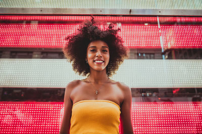 Low angle portrait of happy young woman standing against neon american flag in city
