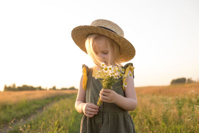 A little blonde girl in a straw hat walks in a field with a bouquet of daisies. 