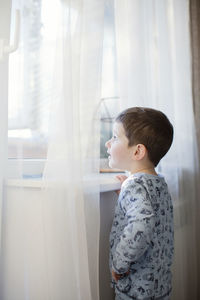 Boy wearing pajamas looking through window at home