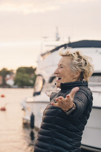 Side view of excited senior woman standing with arms outstretched at harbor