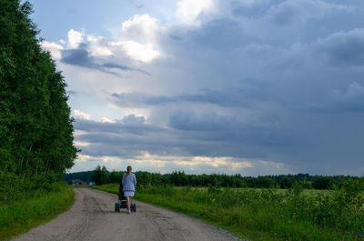 Rear view of woman with baby carriage on dirt road amidst field against sky