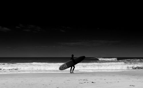 Silhouette person with surfboard standing at beach against sky during sunny day