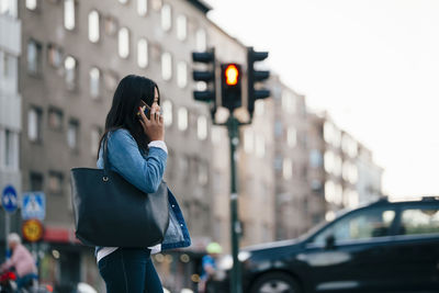 Side view of teenage girl talking on smart phone while walking against building in city