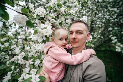 Dad holds a joyful baby daughter in his arms in a garden with a blooming apple tree