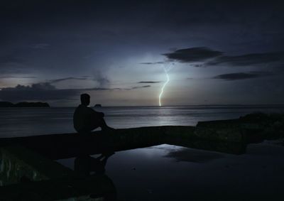 Silhouette man sitting on beach against sky at dusk