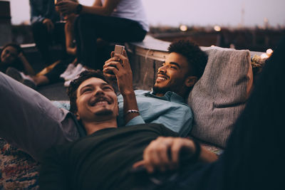 Smiling young man lying while friend using smart phone on terrace at dusk