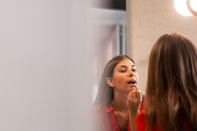 Attractive young woman in casual red outfit applying makeup and using lip pencil while standing in front of mirror at home