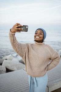 Young woman smiling while taking selfie on mobile phone while standing against sky