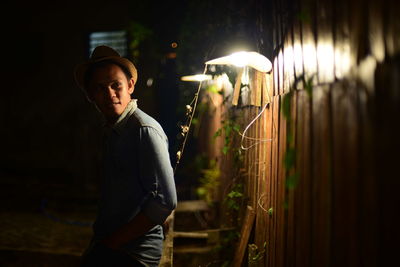 Side view of boy wearing hat standing and looking away by illuminated lamp at night