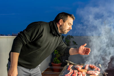 Man preparing food on barbecue grill against sky on terrace