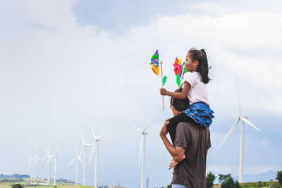 Side view of daughter with pinwheel toys sitting on father shoulders with windmills in background