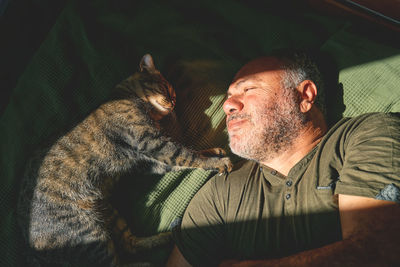 Top view of bearded middle-aged man lying on a bed and playing with his gray tabby cat.