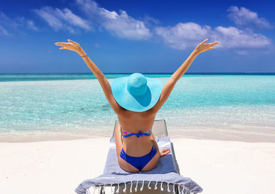 Rear view of woman with arms raised relaxing on chair at beach against sky
