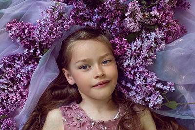 Child girl in a purple floral dress lies on the ground among lilacs on a veil in spring