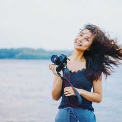 Portrait of young woman holding camera against sky