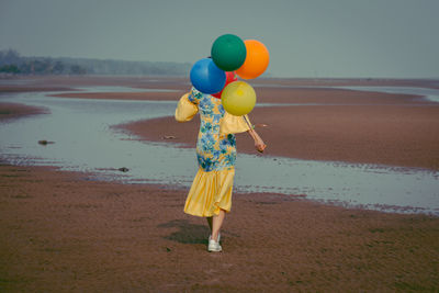 Rear view full length of young woman holding colorful balloons at beach