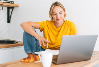 Young smiling blonde woman in yellow t-shirt working at laptop in kitchen at home