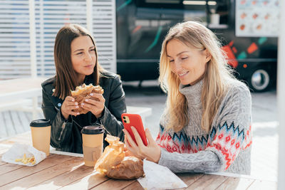 Women eat street fast food and chatting while sitting at a table outside and using phone for video.