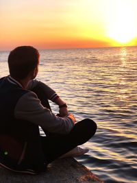 Man sitting by sea against sky during sunset