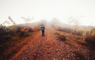 Rear view of man walking on dirt road against clear sky during foggy weather