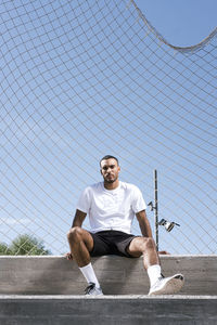 Portrait of man sitting on fence against sky