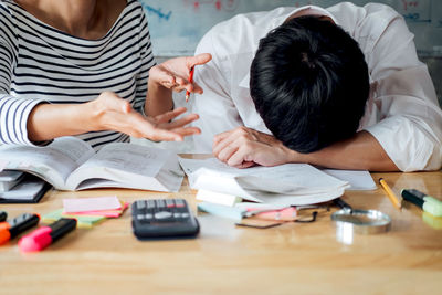 Midsection of woman gesturing by tired man napping by books on desk