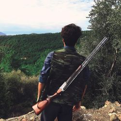 Rear view of young man with rifle standing on rock against sky