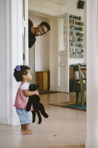 Happy father peeking at daughter holding stuffed toy in house