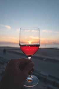 Close-up of hand holding wineglass against sky during sunset