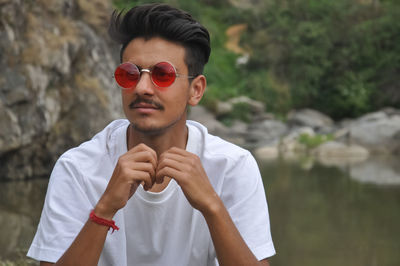 A young guy sitting beside of river with looking sideways  with wearing white t-shirt and sunglasses 