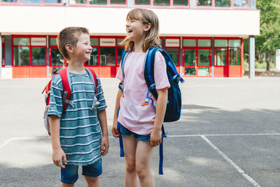 A boy and a girl, brother and sister stand with backpacks on their backs in front of the school 