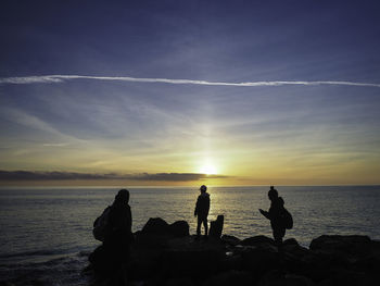 Silhouette people on rocks by sea against sunset sky