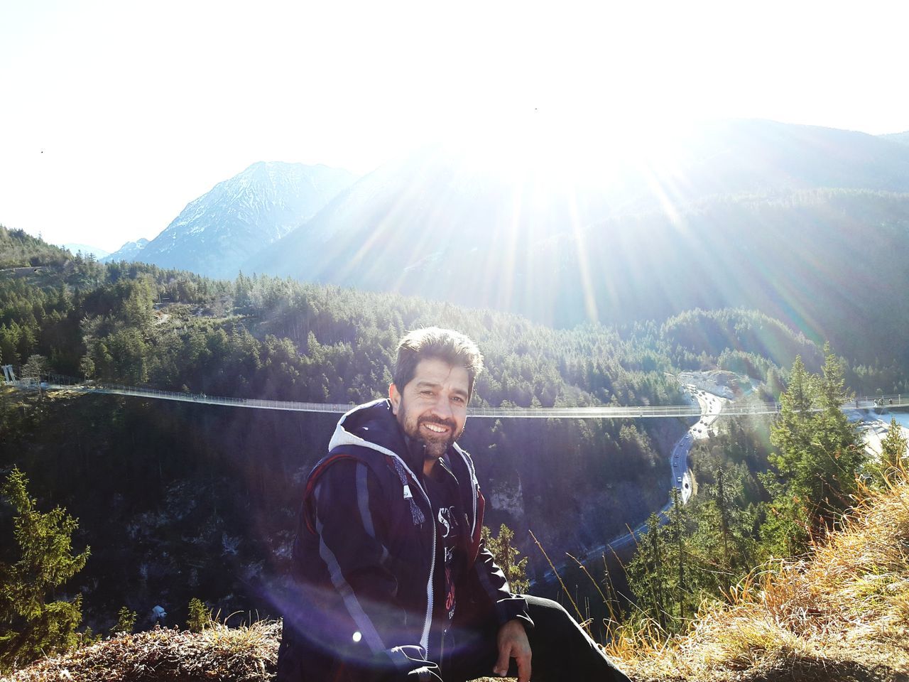 mountain, real people, nature, lens flare, sunlight, one person, leisure activity, looking at camera, beauty in nature, day, happiness, outdoors, lifestyles, smiling, portrait, young adult, scenics, tree, standing, landscape, young women, mountain range, sitting, water, clear sky, sky, people