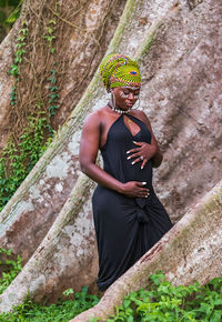 Pregnant african tribal woman standing in the jungle with a large tree trunk root in the background