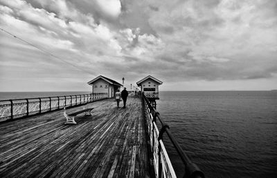 Man and woman walking on bridge over sea against cloudy sky