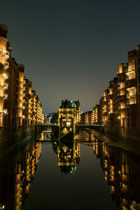 Illuminated buildings by lake against sky in city at night