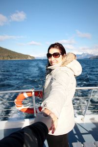 Portrait of smiling woman holding cropped hand on boat sailing in sea against sky