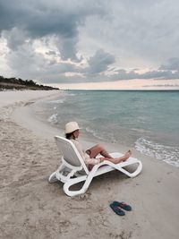 Young woman sitting on lounge chair at beach against sky