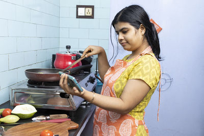 A pretty indian young woman wearing apron watching cooking video in smart phone in domestic kitchen