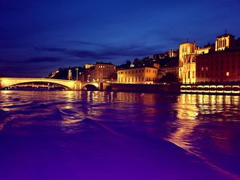 Illuminated bridge over river by buildings against sky at night