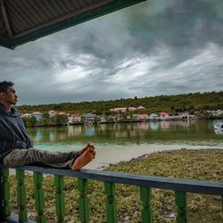 Man sitting on railing by lake against sky