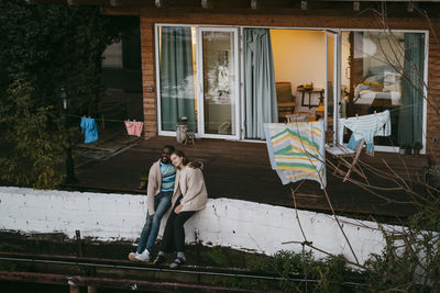 Multiracial couple enjoying with each other while sitting on retaining wall outside house