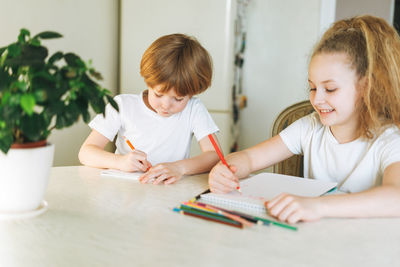 Two siblings brother and sister toddler boy tween girl drawing on table in kitchen at the home