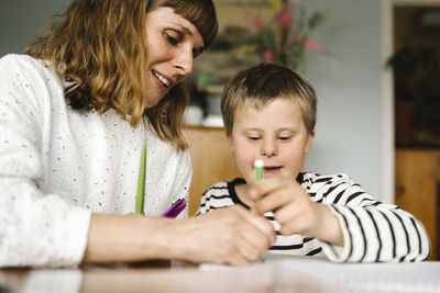 Smiling mother teaching son with disability to draw in book at home