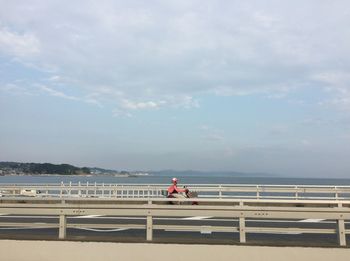 Man sitting on railing by sea against sky