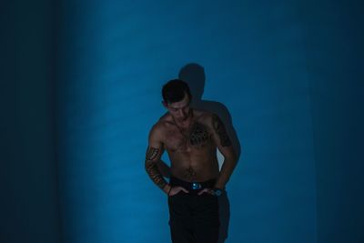 Shirtless tattooed man with hands in pockets standing against blue wall