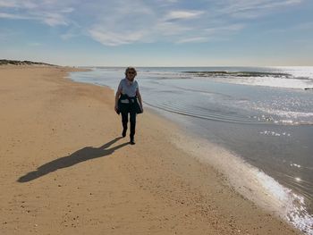 Full length of senior woman walking on beach against sky during sunny day