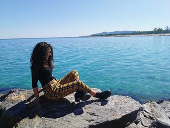 Side view of woman sitting on rock by sea against clear sky during summer