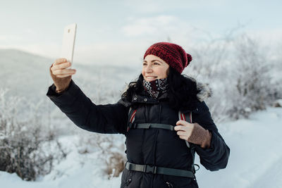 Smiling woman in warm taking selfie while standing on snowcapped mountain